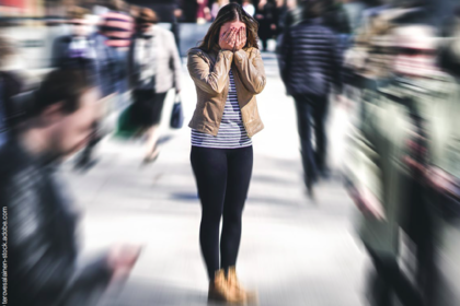 Vor einem Hintergrund mit Bewegungsunschärfe vieler undeutlicher Menschen in einem Fußgängerzonensetting eine junge Frau mit offenen dunklen Haaren, die sich die Hände vor ihr Gesicht hält. Sie träge eine beige Jacke über weißem Shirt mit dunklen Querstreifen sowie schwarze Hose und beige Schuhe.