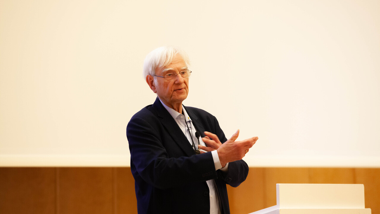 Prof. Dr. Dr. h. c. Dieter Birnbacher steht, bekleidet mit dunkelblauen Anzug und hellblaues Hemd vor dunkler Holzvertäfelung und heller Projektionsfläche hinter einem hellen Pult. Ein trägt eine Brille und ein umgehängtes Funkmikrofon, vor dem er mit den Händen gestikuliert. Deutet mit der rechten Hand Richtung Publikum und hält die linke Hand über die eigene Brust.
