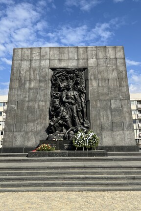 Bronzerelief des Warschauer Ghetto-Ehrenmals von Natan Rapoport in Warschau mit mehreren kämpferisch dargestellten Figuren; am Sockel liegt ein blau-weiß geschmückter Gedenkkranz.