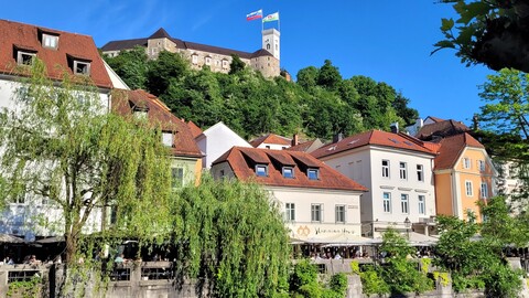 Blick auf einen Hügel mit bunten Gebäuden mit roten Dächern und einer üppigen Kulisse. Auf dem Hügel steht unter einem klaren blauen Himmel eine Burg, auf der zwei Fahnen wehen.