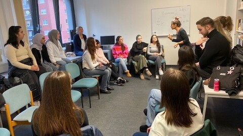 Lehrende Person steht vor einem Whiteboard und erklärt etwas. Lernende Personen hören aufmerksam zu.