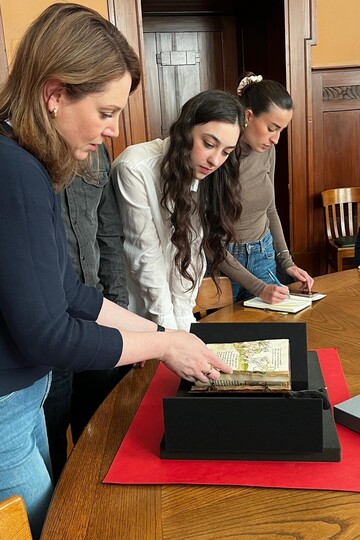 Ein Paar Hände blättert in einer spätmittelalterlichen Handschrift, das auf einer Schaumstoffunterlage liegt. Im Hintergrund macht sich jemand Notizen.