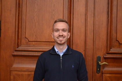 Maximilian Spanier vor der Eingangstür des Mittelbaus der Universität Mannheim. Er trägt einen dunkelblauen Pullover, darunter ein hellblaues Hemd und lächelt. 