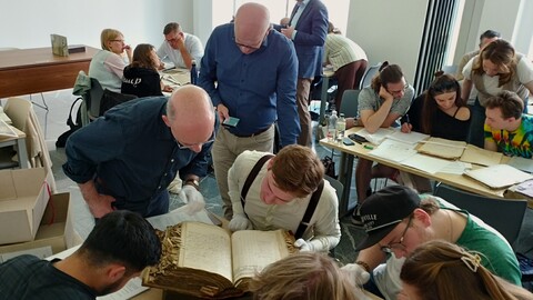Die Teilnehmenden der Summer School diskutieren in Kleingruppen lebhaft und studiert gemeinsam historische Handschriften in einem Klassenzimmer.