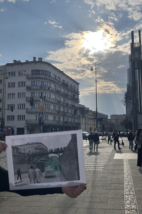 Eine Straßenkreuzung in Warschau. Im Hintergrund ein großes Gebäude an der Kreuzung. Im Vordergrund wird eine historische Fotografie hochgehalten, auf welcher das gleiche Eckhaus abgebildet ist, wobei auf dem historischen Foto ebenfalls Ghettomauern und eine Brücke zu sehen sind. 