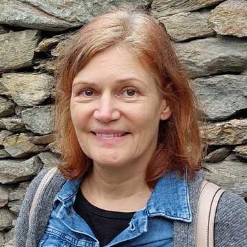 Jutta Linz hat rötliches, schulterlanges Haar und lächelt, im Hintergrund eine Steinmauer. 