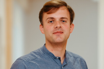 Mann mit braunem Haar trägt ein blaues Hemd mit Knöpfen, steht in einem hellen Flur