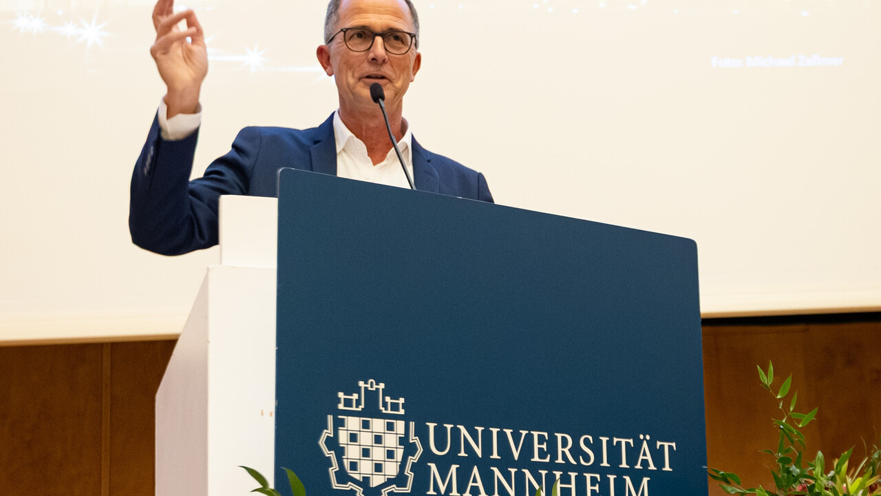Der Prodekan für Forschung Prof. Müller-Lancé steht am mit Blumen dekorierten Rednerpult und betont seine Moderation mit einer Handgeste.