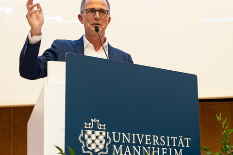 Der Prodekan für Forschung Prof. Müller-Lancé steht am mit Blumen dekorierten Rednerpult und betont seine Moderation mit einer Handgeste.