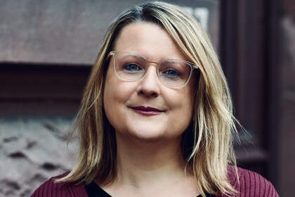 Maria Alexopoulou trägt ein rotes Oberteil. Sie hat schulterlanges blondes Haar und eine Brille. 