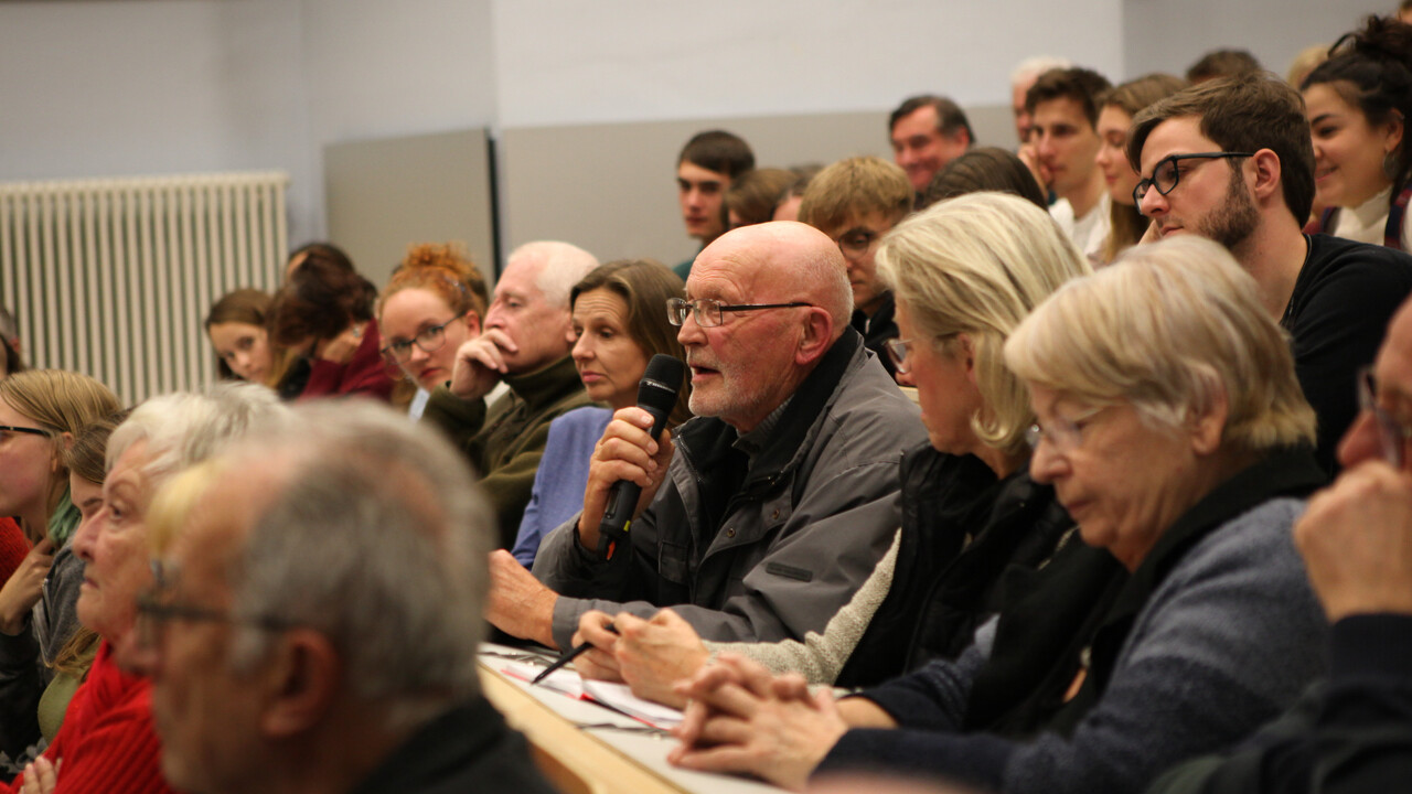 Zuschauerfrage: Inmitten anderer Vortragsgäste auf den vollbesetzten Hörsaalsitzen hält ein mit grauer Jacke gekleideter älterer Mann mit Brille ein Mikrofon in der Hand und spricht hinein.