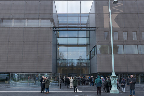 Viele Menschen stehen vor einem Gebäude. Das Gebäude hat eine braune Fassade und viele Fenster.
