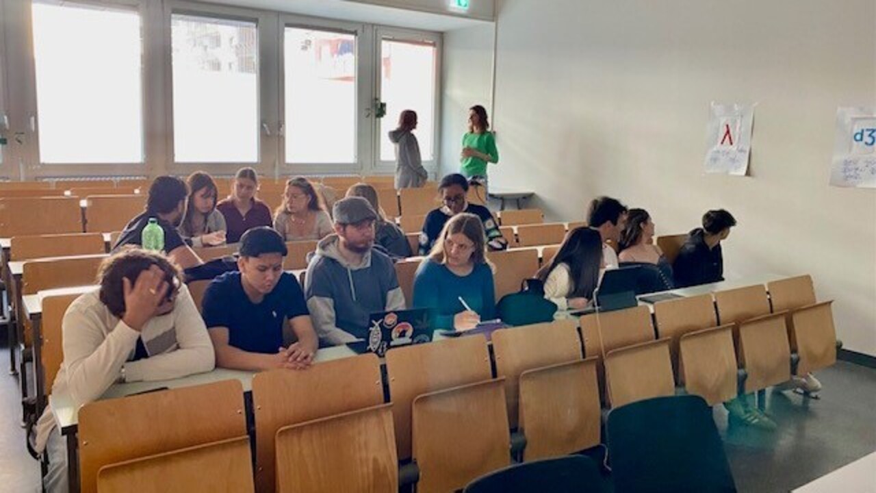 Schüler*innen eines Gymnasiums und Studierende sitzen in einem Hörsaal und arbeiten gemeinsam an einer Aufgabe.