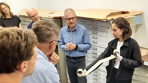 Die Teilnehmenden der Summer School stehen in einem Depotraum in einem Archiv. Eine Frau hält eine aufgerollte Pergamentrolle in der Hand, während andere ihr aufmerksam zuhören.