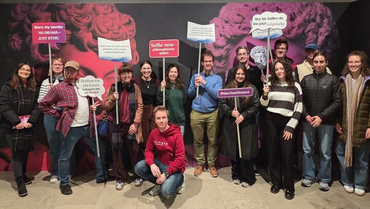 Die Teilnehmenden der Exkursion in der Ausstellung „Marc Aurel - Kaiser, Feldherr, Philosoph“ im Rheinischen Landesmuseum. Die Teilnehmenden stehen vor einer Fotowand und halten Schilder hoch, darauf beispielsweise die Sprüche "Marc my words: NO DRAMA - JUST STOIC", "Selfie now-philosophieren später", "#MarcYourMoment" und "Denken wie ein Kaiser. schauen wie ein Model!".  