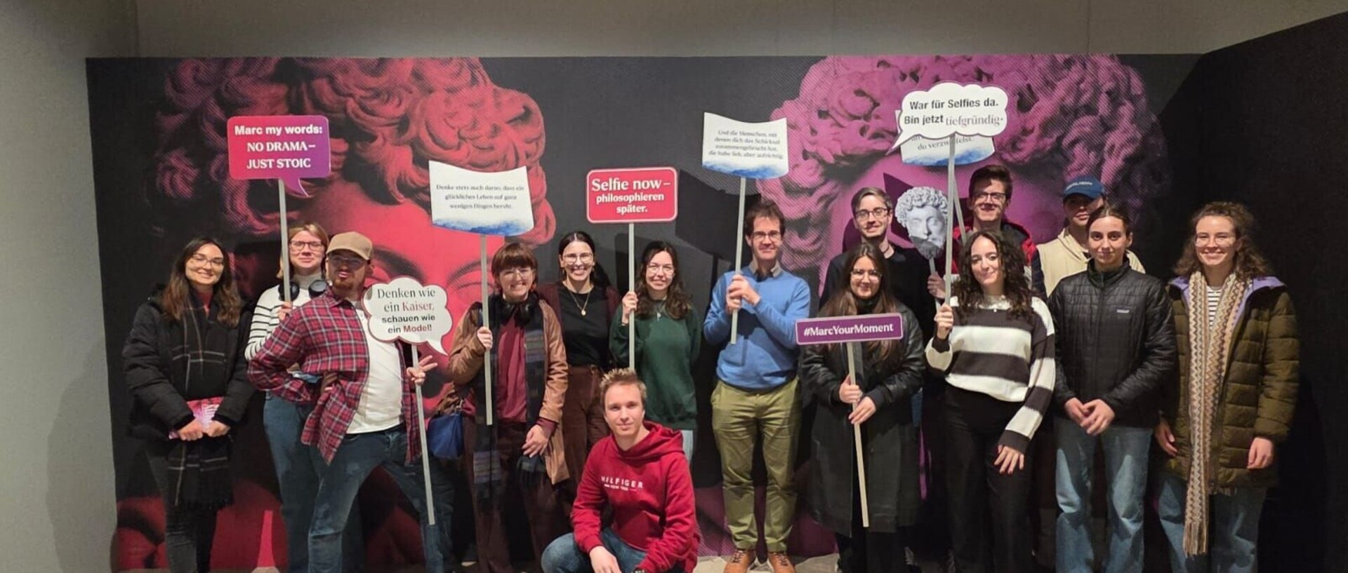 Die Teilnehmenden der Exkursion in der Ausstellung „Marc Aurel - Kaiser, Feldherr, Philosoph“ im Rheinischen Landesmuseum. Die Teilnehmenden stehen vor einer Fotowand und halten Schilder hoch, darauf beispielsweise die Sprüche "Marc my words: NO DRAMA - JUST STOIC", "Selfie now-philosophieren später", "#MarcYourMoment" und "Denken wie ein Kaiser. schauen wie ein Model!".  