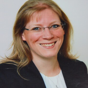 Birgit Füreder hat blonde Haare. Sie trägt einen schwarzen Blazer, ein weißes T-Shirt und eine Brille.