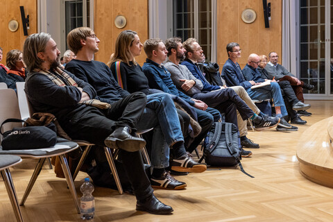 Seitlicher Blick von Vorne auf die erste Reihe des Publikums am Tag der Philosophie 2025 in Mannheim: Im Bild sind etwa ein Dutzend herbstlich bekleidete, auf Stühlen sitzende, Personen zu sehen. Das Bild ist geprägt vom hölzernen Parkettboden sowie rechts im Anschnitt sichtbarem Bühnenaufgang und der Wandvertäfelung der Aula, welche im Hintergrund von hohen Fenstern mit weißem Anstrich ihrer Einfassungen durchbrochen wird.