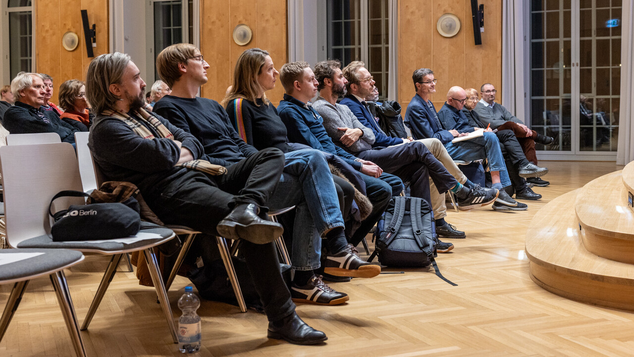 Blick auf das Publikum in der Aula Seitlicher Blick von Vorne auf die erste Reihe des Publikums am Tag der Philosophie 2025 in Mannheim: Im Bild sind etwa ein Dutzend herbstlich bekleidete, auf Stühlen sitzende, Personen zu sehen. Das Bild ist geprägt vom hölzernen Parkettboden sowie rechts im Anschnitt sichtbarem Bühnenaufgang und der Wandvertäfelung der Aula, welche im Hintergrund von hohen Fenstern mit weißem Anstrich ihrer Einfassungen durchbrochen wird.
