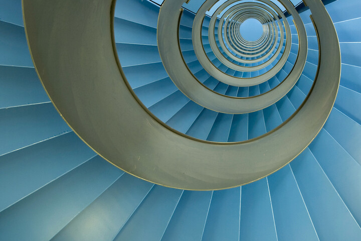 Wendeltreppe mit endlosen blauen Facetten. Präziser: Mit mindestens 12 Rundungen sich in Blickrichtung verjüngende Spirale aus blauen und goldenen Elementen. Die blauen Elemente sind in Rechtecke mit Farbverlauf bzw. -nuancen ins Weiße unterteilt, das goldene Element läuft dagegen fast fortlaufend und ist nur durch 3 Einkerbungen unterbrochen, die jeweils Anfang, Mitte und Ende einer Umrundung bilden. Bei genauer Betrachtung bemerkt man, dass sich im blauen Bereich 20 eher kleine und dunkle mit 5 hellen und großen Elementen abwechseln. Ein im Vergleich zu den kleineren noch einmal nur etwa halb so breites letztes Element ist dunkelblau bis schwarz und bildet den Abschluss bevor sich dieses Muster wiederholt. Die gennanten Absetzungen bilden in ihrer Gänze mehrere durchbrochene Linien die alle dem Mittelpunkt der sich optisch verjüngenden Spirale zustreben.