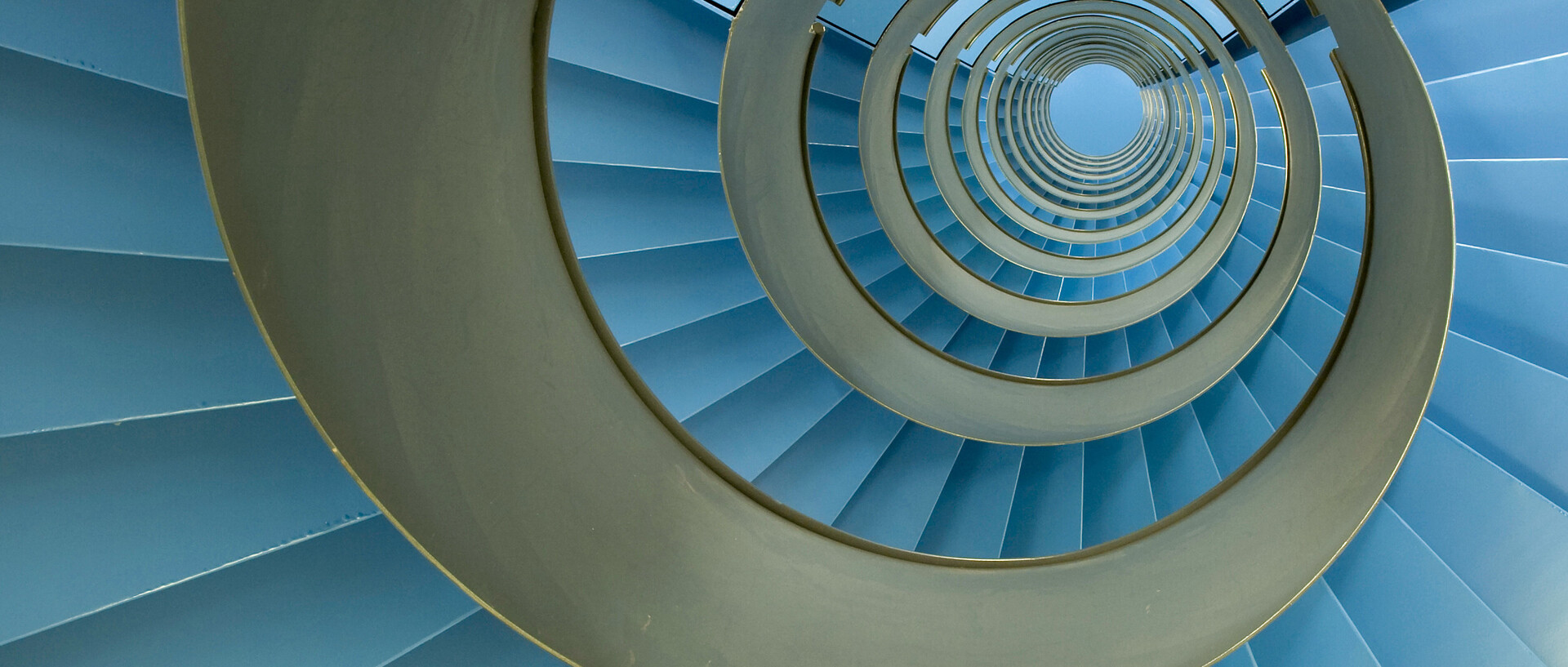 Spiral staircase with endless blue facets. More precisely: A spiral of blue and gold elements tapering in the direction of view, with at least 12 curves. The blue elements are divided into rectangles with color gradients or shades of white, while the gold element runs almost continuously and is interrupted only by three indentations, each forming the beginning, middle, and end of a circle. On closer inspection, one notices that in the blue area, 20 rather small and dark elements alternate with 5 light and large elements. The last element, which is only about half as wide as the smaller ones, is dark blue to black and forms the end before this pattern repeats itself. Together, these indentations form several broken lines that all converge toward the center of the visually tapering spiral.