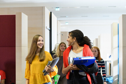 Eine Gruppe weiblicher Studierender mit Lernsachen in den Händen läuft durch die Bibliothek und unterhält sich