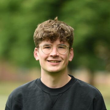 Jakob Grosch trägt einen schwarzen Pullover. Er hat kurze braune Haare, trägt eine silberne Brille und lächelt. 