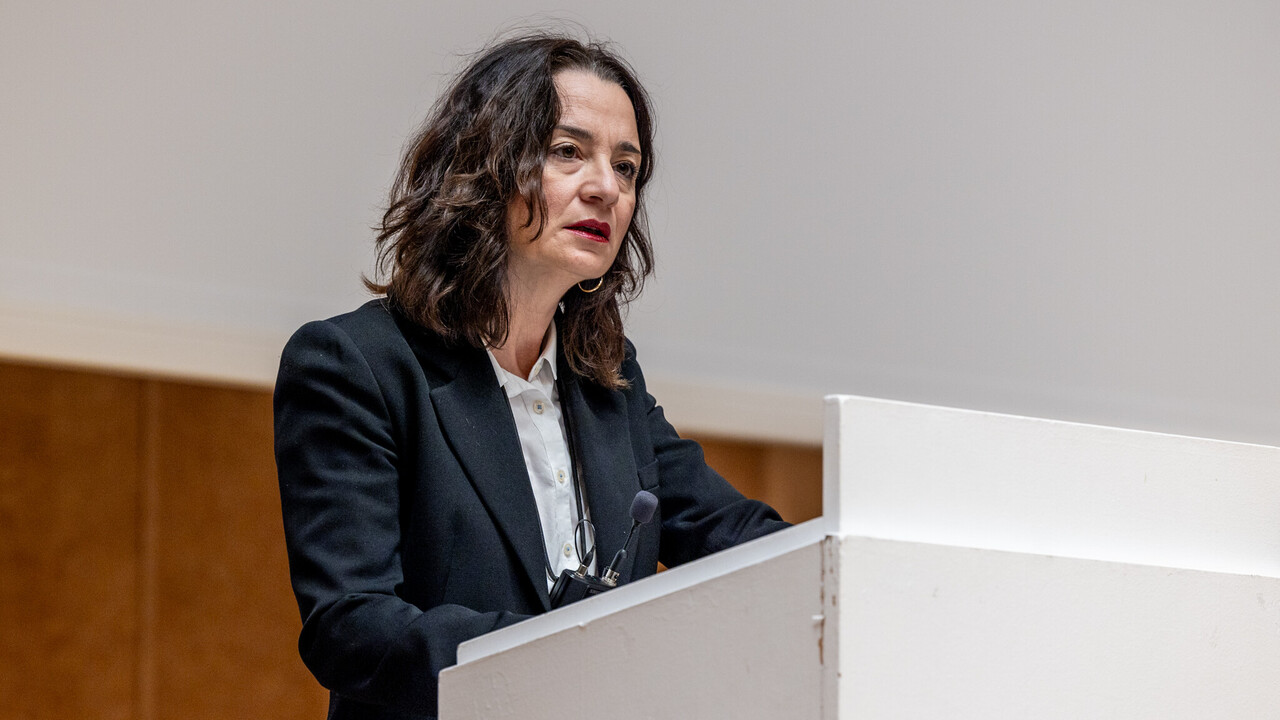 Vortrag durch Prof. Dr. phil. Elif Özmen Prof. Dr. phil. Elif Özmen mit umgehängtem Funkmikrofon hinter dem hellen Rednerpult beim Vortrag zum Tag der Philosophie am 20.11.2025. Sie trägt ein helles Hemd und eine dunkle Anzugjacke. Im Hintergrund Holzvertäfelung und Leinwand der Bühne in der Aula.