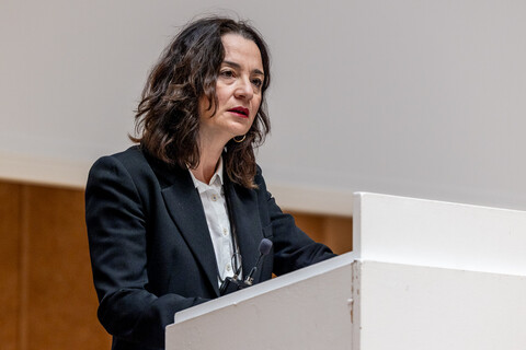 Prof. Dr. phil. Elif Özmen mit umgehängtem Funkmikrofon hinter dem hellen Rednerpult beim Vortrag zum Tag der Philosophie am 20.11.2025. Sie trägt ein helles Hemd und eine dunkle Anzugjacke. Im Hintergrund Holzvertäfelung und Leinwand der Bühne in der Aula.