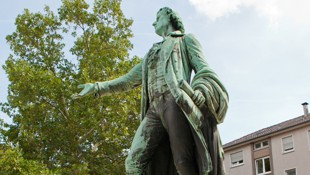 Eine Statue von Friedrich Schiller aus Metall. Die Figur ist grün verfärbt und steht auf grauen Steinen.