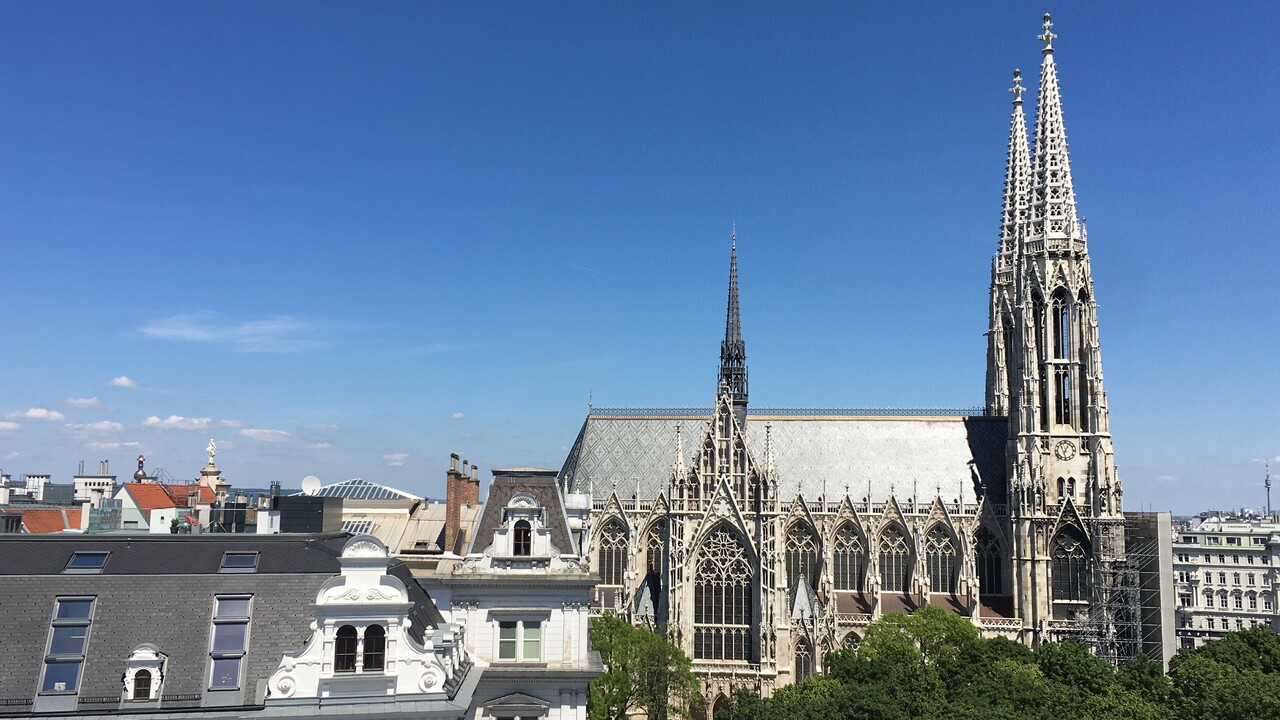 Foto von einem erhöhten Punkt auf Fassaden und Dächer. Die rechte Bildhälfte ist von der Votivkirche in Wien geprägt, vor der grüne Laubbaumkronen zu sehen sind. Am vorderen Turm rechts ist im unteren Bereich ein Baugerüst angebracht.