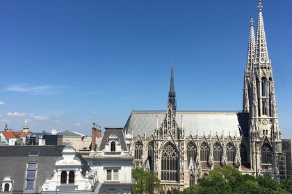 Foto von einem erhöhten Punkt auf Fassaden und Dächer. Die rechte Bildhälfte ist von der Votivkirche in Wien geprägt, vor der grüne Laubbaumkronen zu sehen sind. Am vorderen Turm rechts ist im unteren Bereich ein Baugerüst angebracht.